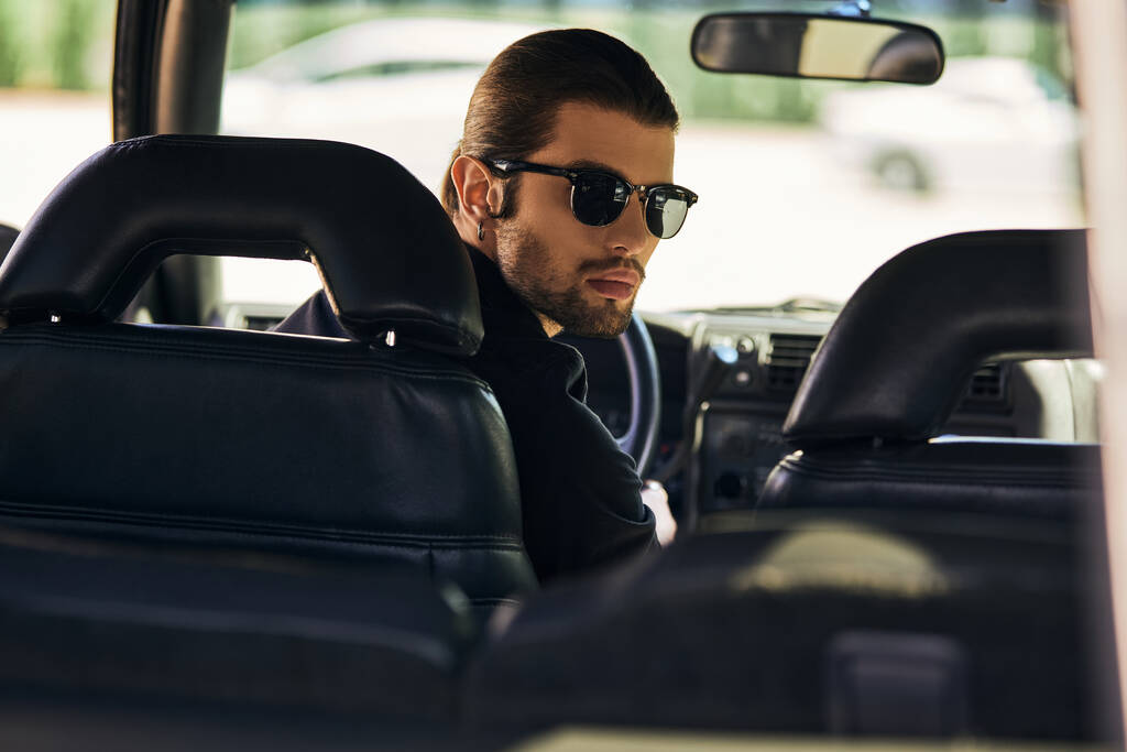 conductor atractivo en traje negro detrás del volante y mirando a la cámara, concepto de moda - Foto, Imagen