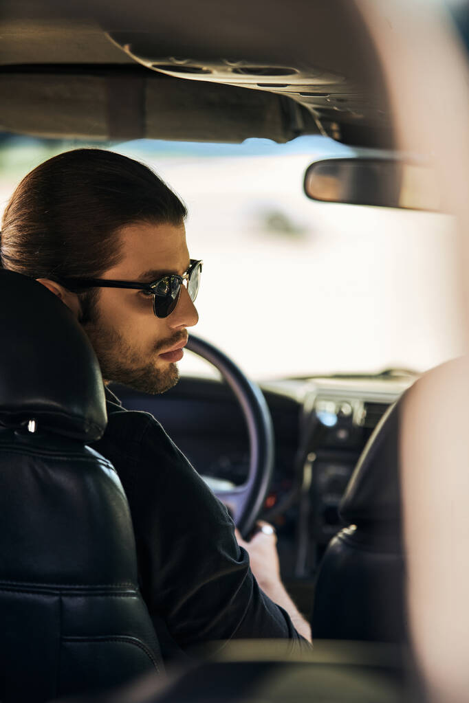 pionowe ujęcie dobrze wyglądającego mężczyzny w czarnym eleganckim stroju przy kierownicy, obracającego głową - Zdjęcie, obraz