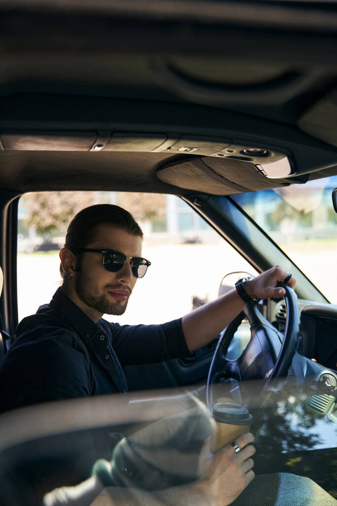 pionowe ujęcie atrakcyjnego eleganckiego mężczyzny w okularach przeciwsłonecznych i zegarku na rękę przy kierownicy z kawą - Zdjęcie, obraz