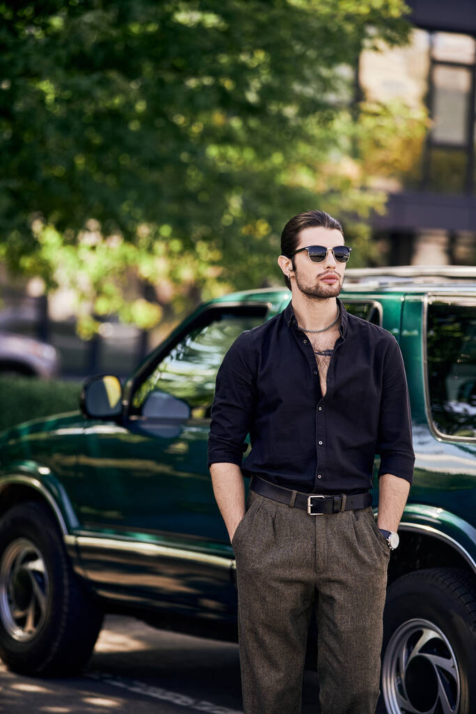 pionowe ujęcie czarującego mężczyzny w eleganckim czarnym stroju, pozującego obok samochodu z rękami w kieszeniach - Zdjęcie, obraz