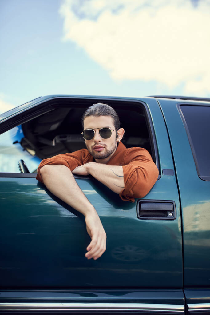 encantador joven en camisa marrón con gafas de sol mirando a través de la ventana del coche, el estilo y la moda - Foto, Imagen