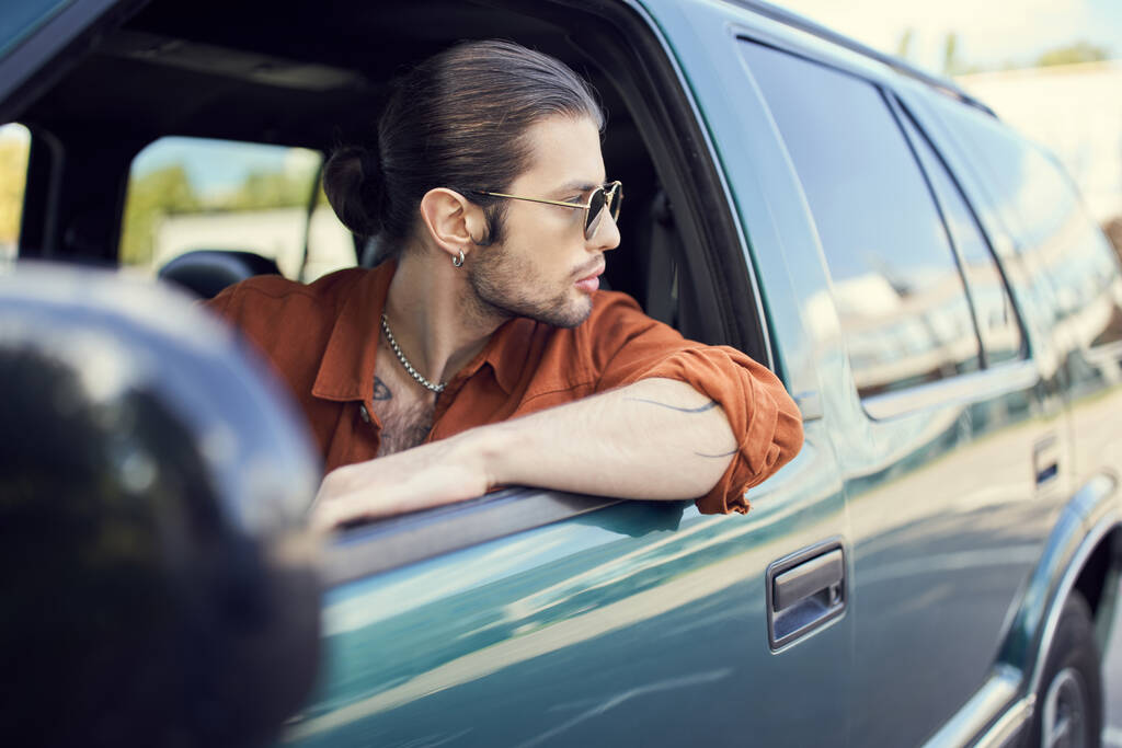 όμορφος άνδρας σε κομψό ζωντανό ντύσιμο με γυαλιά ηλίου και αλογοουρά κοιτάζοντας έξω από το παράθυρο του αυτοκινήτου, στυλ - Φωτογραφία, εικόνα