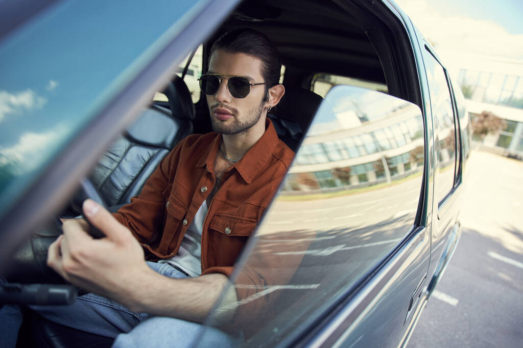 guapo joven modelo masculino en elegante camisa marrón con gafas de sol en el volante, conductor sexy - Foto, imagen