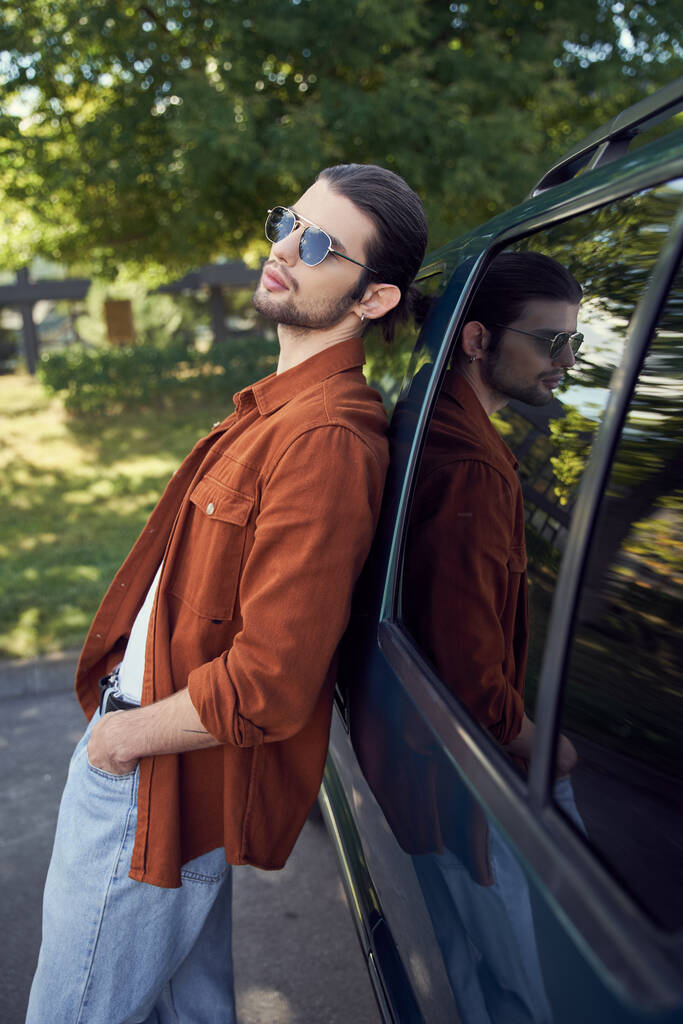 verleidelijke stijlvolle man met een zonnebril poseren buiten leunend licht op zijn auto, mode concept - Foto, afbeelding