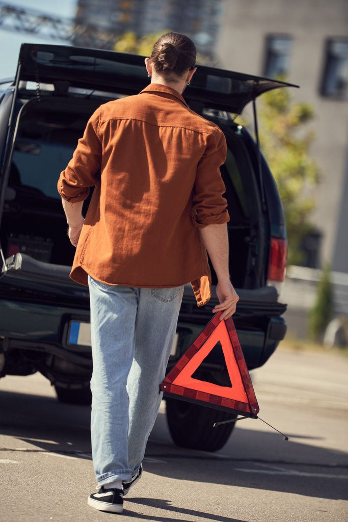 πίσω όψη του νεαρού κομψό άνδρα σε αστική ενδυμασία περπάτημα στο αυτοκίνητό του με προειδοποιητικό τρίγωνο στα χέρια - Φωτογραφία, εικόνα