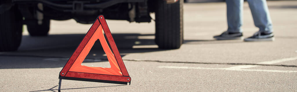 обрезанный вид на предупреждающий треугольник перед молодым мужчиной-моделью, стоящим возле его машины, баннер - Фото, изображение