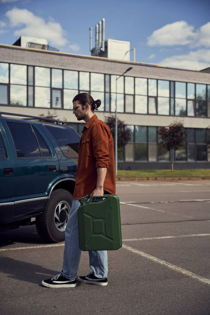όμορφος νεαρός άνδρας σε καφέ πουκάμισο με τζιν με κάνιστρο βενζίνης στο χέρι με τα πόδια στο αυτοκίνητό του - Φωτογραφία, εικόνα