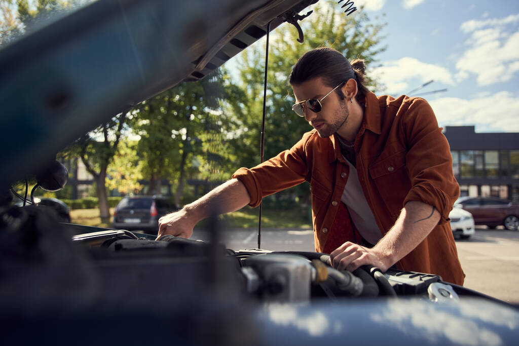νεαρός άνδρας με γυαλιά ηλίου και γενειάδα σε καφέ κομψό πουκάμισο και τζιν έλεγχο στη μηχανή του αυτοκινήτου του - Φωτογραφία, εικόνα