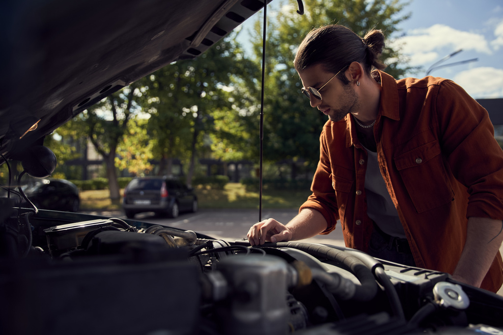 pensive handsome man in stylish outfit with sunglasses and ponytail checking on his car engine - Photo, Image