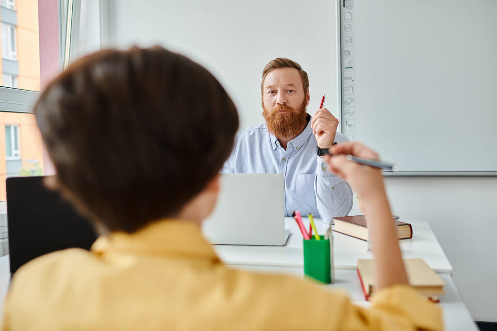 Een man met een baard zit aan een bureau, bezig met denken, mogelijk een les voorbereiden of onderzoek doen.. - Foto, afbeelding