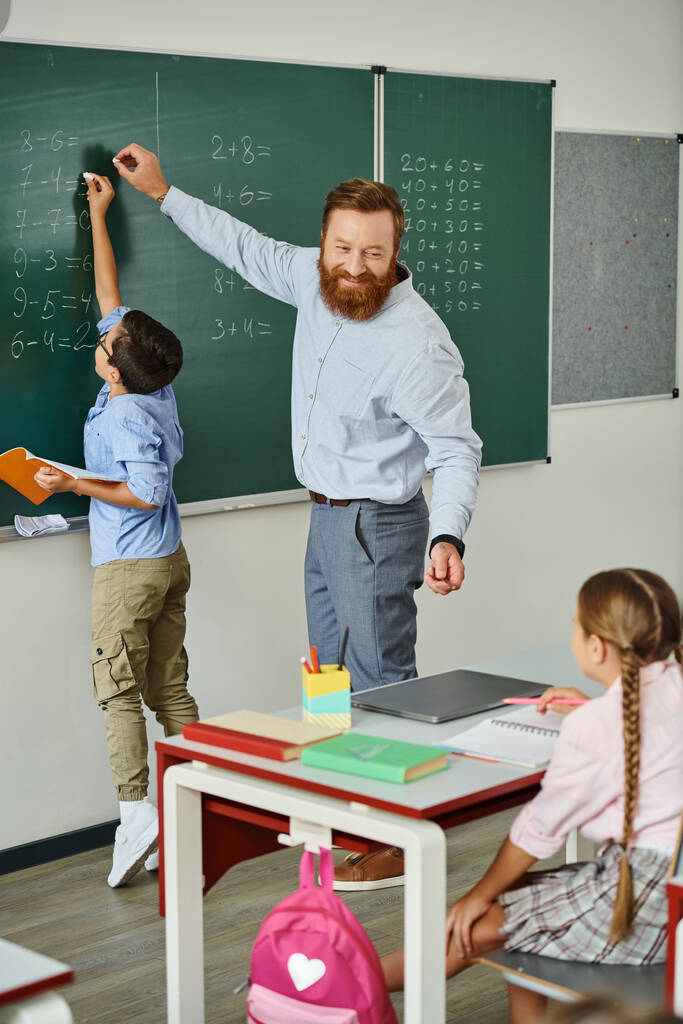 Samiec nauczyciela stoi pewnie przed tablicą, z pasją kształcąc grupę dzieci w jasnym, żywym otoczeniu szkolnym.. - Zdjęcie, obraz