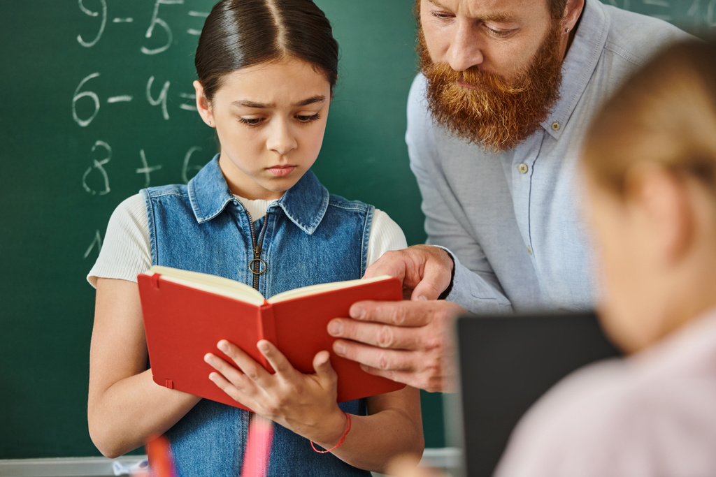 Egy férfi tanár könyvet olvas egy fiatal lánynak, aki érdeklődik egy élénk osztálytermi környezetben. - Fotó, kép