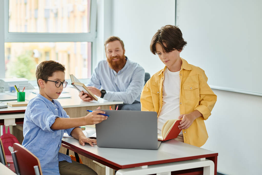 Ein kleiner Junge sitzt aufmerksam an einem Laptop, während ein Lehrer in einem hellen und lebhaften Klassenzimmer genau beobachtet. - Foto, Bild
