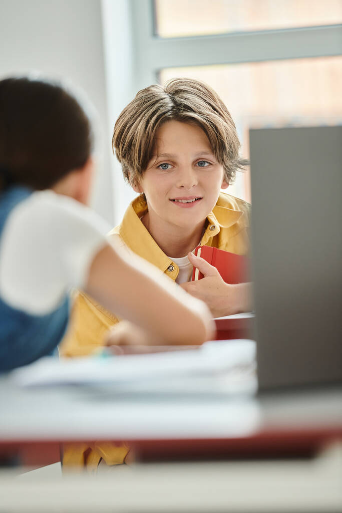 Un jeune garçon assis à un bureau dans une salle de classe dynamique, absorbé dans son écran d'ordinateur portable - Photo, image