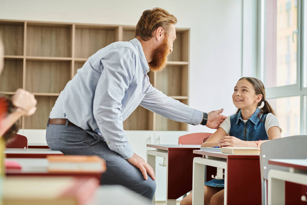 Ein männlicher Lehrer ermutigt ein kleines Mädchen in einem bunten, energiegeladenen Klassenzimmer. - Foto, Bild