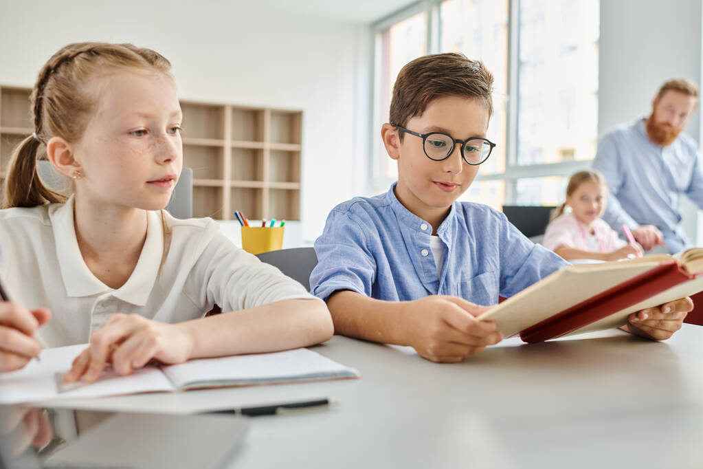 Een jongen en een meisje zitten aan een tafel, verdiept in het lezen van een boek samen in een heldere klas setting. - Foto, afbeelding