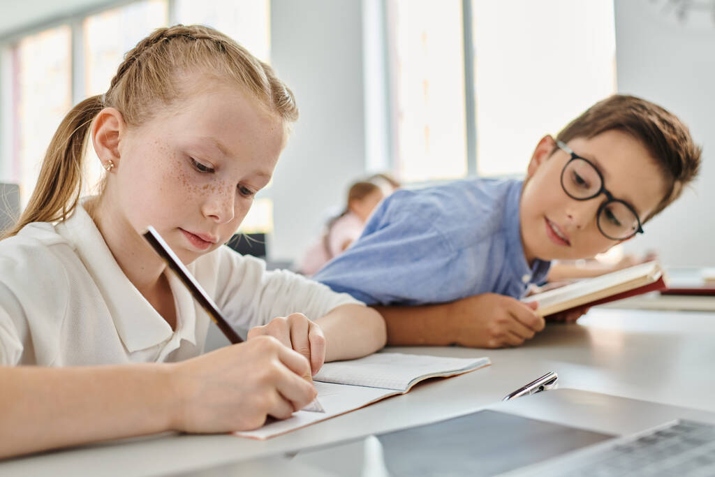 Ein junges Mädchen und ein Junge voller Gedanken, während sie in einem lebhaften Klassenzimmer ihre Hausaufgaben erledigen. - Foto, Bild