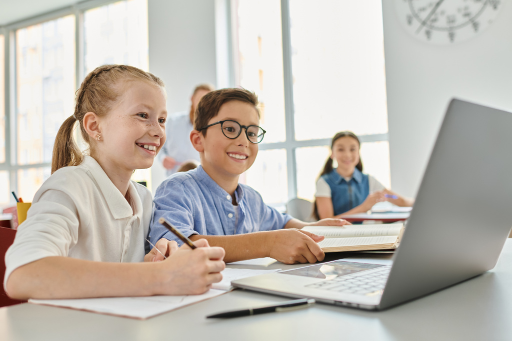 Група дітей, що сидять за столом, зосереджена на ноутбуці в яскравому класі. - Фото, зображення