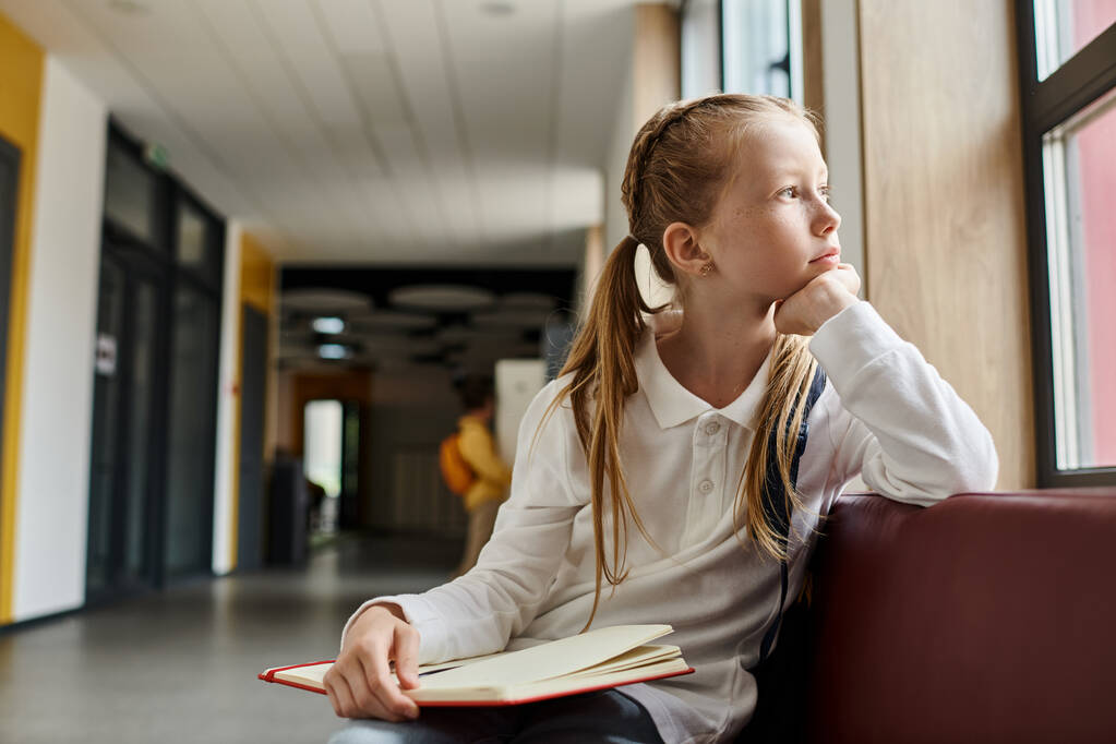 Ein junges Mädchen mit langen dunklen Haaren sitzt allein auf einer Holzbank, in ein Buch vertieft, tief in Gedanken. - Foto, Bild