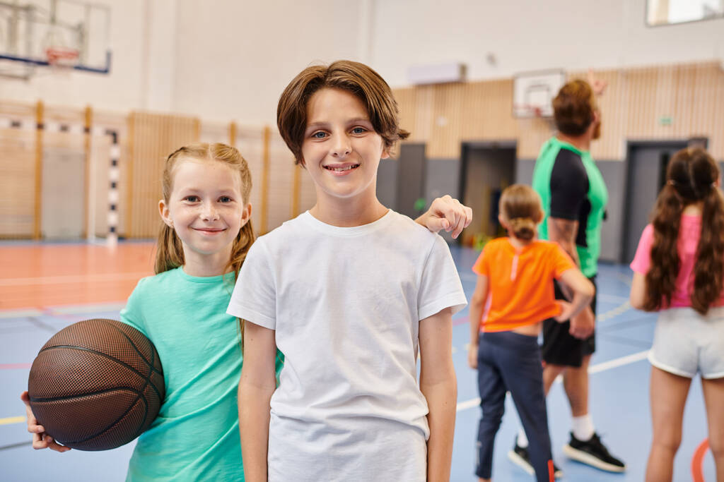 Dwoje dzieci, jeden chłopiec i dziewczyna, stoją obok siebie, trzymając koszykówkę i uśmiechając się szczęśliwie. - Zdjęcie, obraz