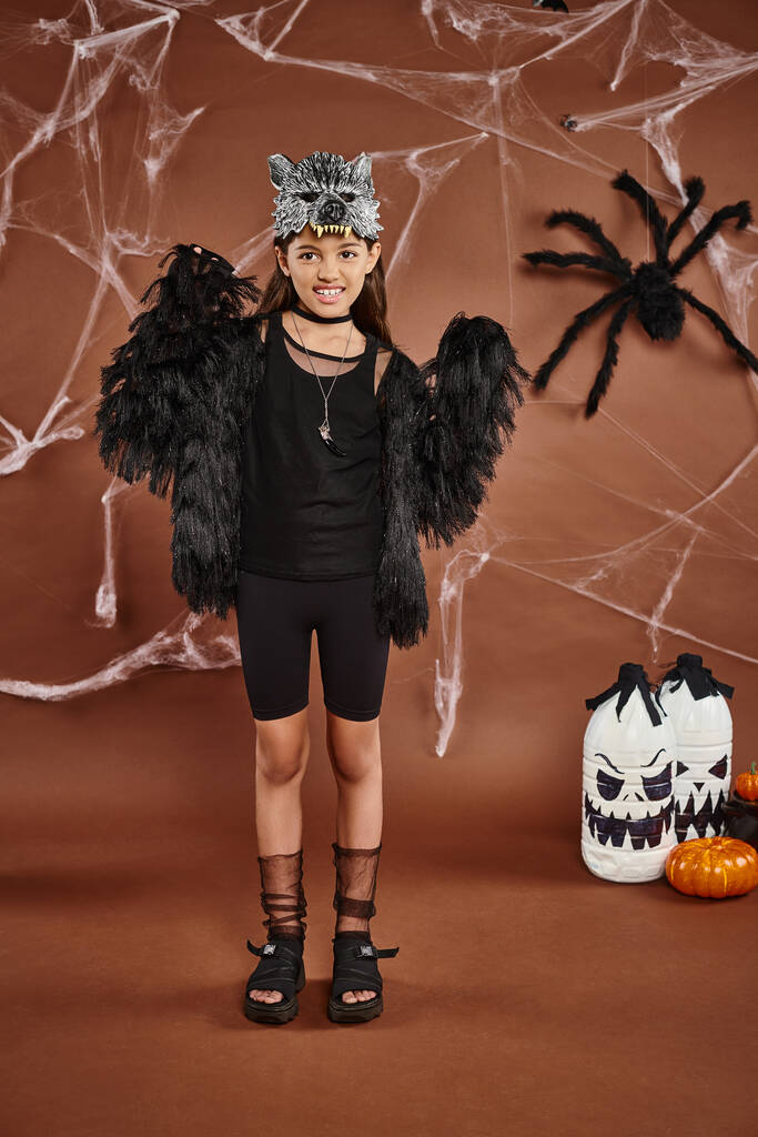 Nettes Mädchen in schwarzer Kleidung und Maske, das zwischen Spinnen, Laternen und Spinnweben Angst und Schrecken verbreitet, Halloween-Konzept - Foto, Bild