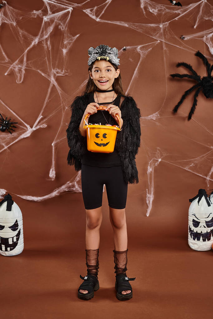 happy girl holding bucket of sweets in wolf mask and black attire, Halloween concept - Photo, Image