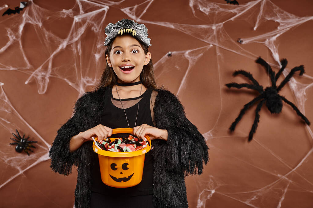 close-up vrolijk meisje met haar emmer vol snoep op bruine achtergrond, Halloween concept - Foto, afbeelding