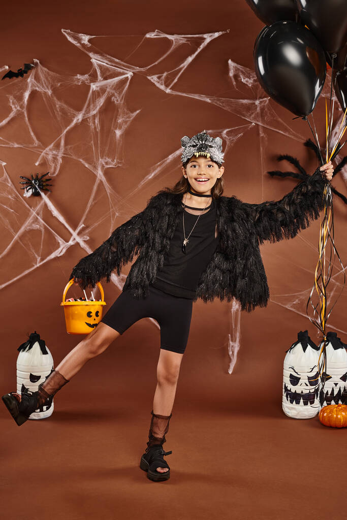 fille heureuse debout sur une jambe tient ballons noirs et orange et seau avec des bonbons, Halloween - Photo, image