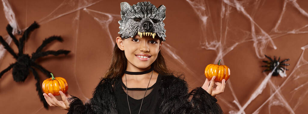 tiener meisje in wolf masker houdt pompoenen op bruine achtergrond met spinnen en web, Halloween, banner - Foto, afbeelding