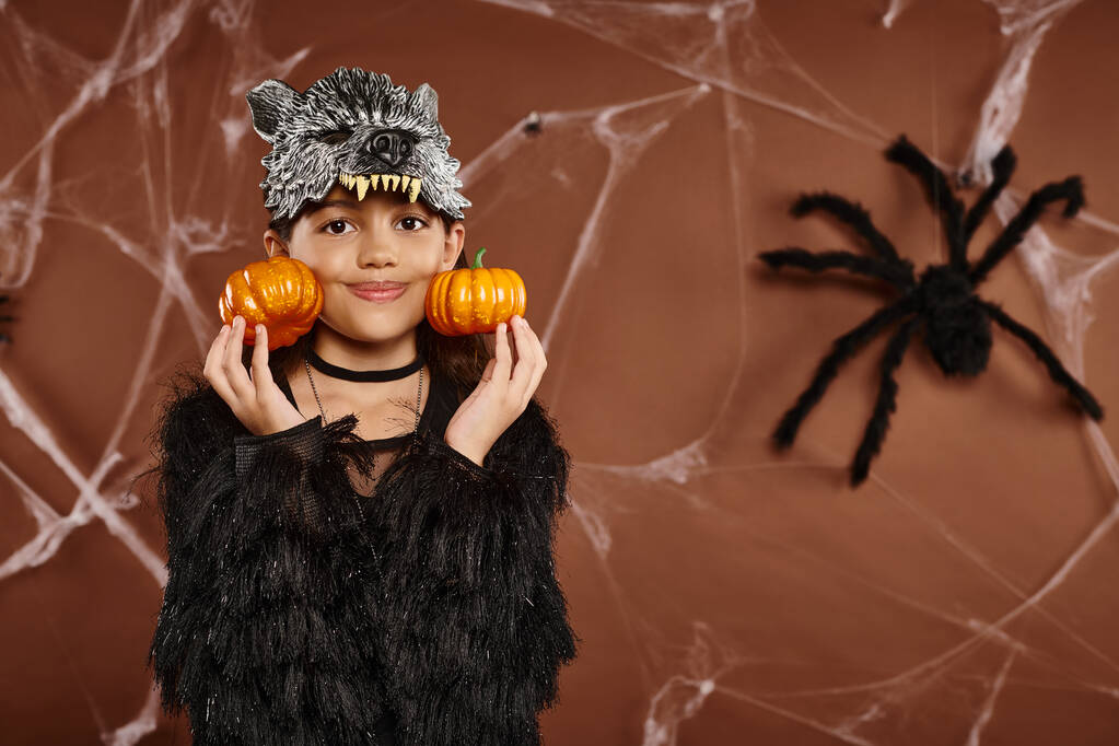 cerca de niña sonriente en máscara de lobo sostiene calabazas cerca de su cara con araña en el telón de fondo, Halloween - Foto, Imagen
