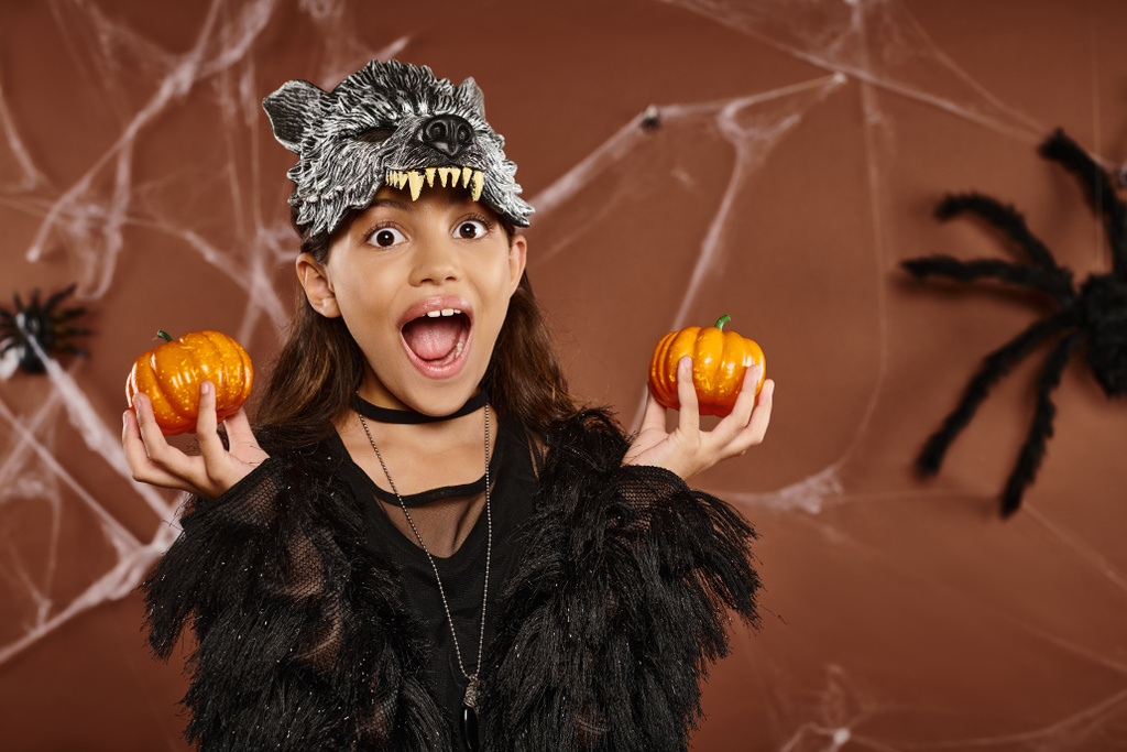 κοντινό ενθουσιασμένο κορίτσι με μάσκα λύκου κρατά κολοκύθες στα χέρια της με αράχνη στο φόντο, Απόκριες - Φωτογραφία, εικόνα