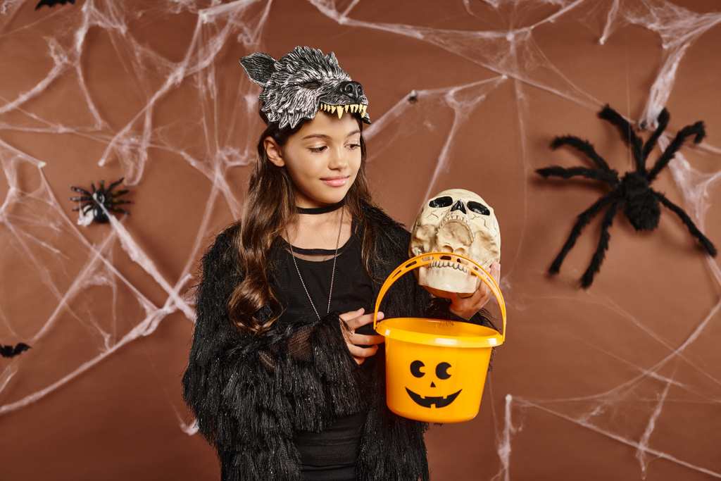 lächelndes Mädchen mit Totenkopf und Eimer voller Süßigkeiten, brauner Hintergrund mit Spinnweben, Halloween - Foto, Bild