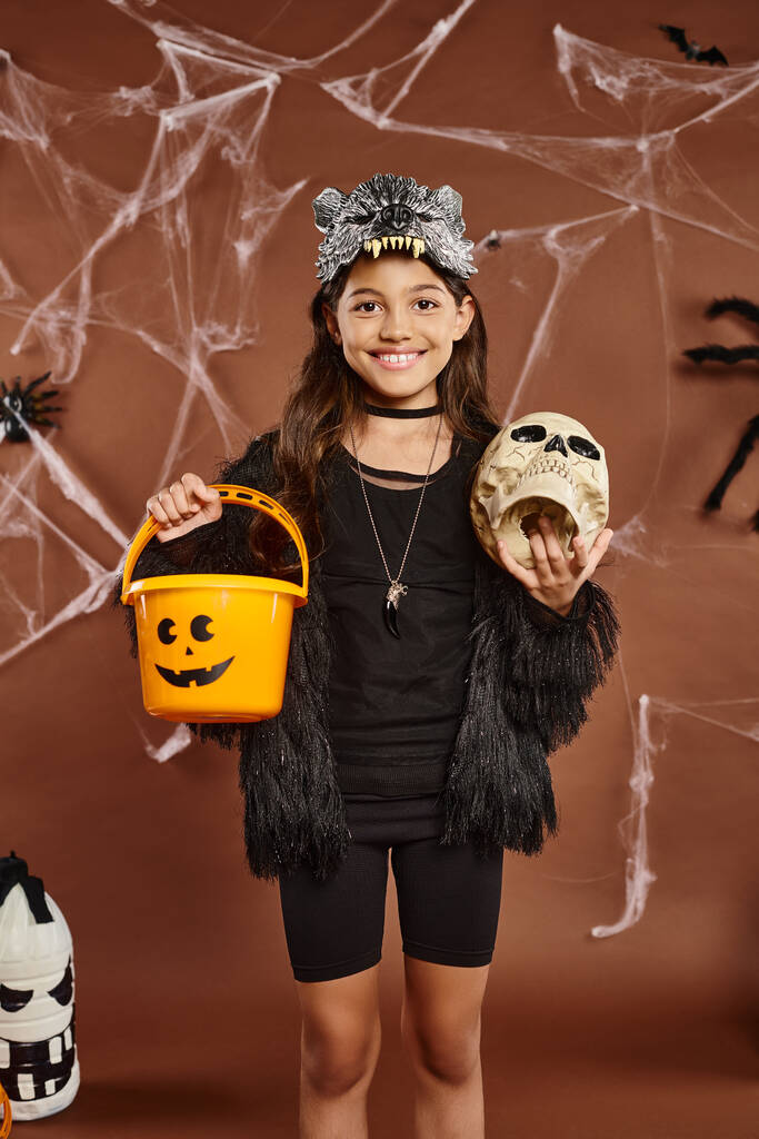 Nahaufnahme lächelndes Mädchen mit Totenkopf und Eimer voller Süßigkeiten, brauner Hintergrund mit Netz, Halloween - Foto, Bild