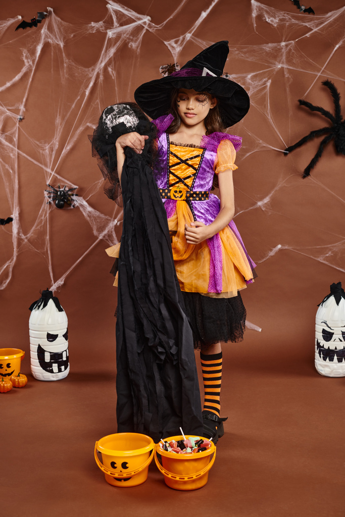 Smiley-Mädchen mit Hexenhut und Spinnennetz-Make-up mit gruseligem Spielzeug in der Nähe von Kürbiseimern, Halloween - Foto, Bild