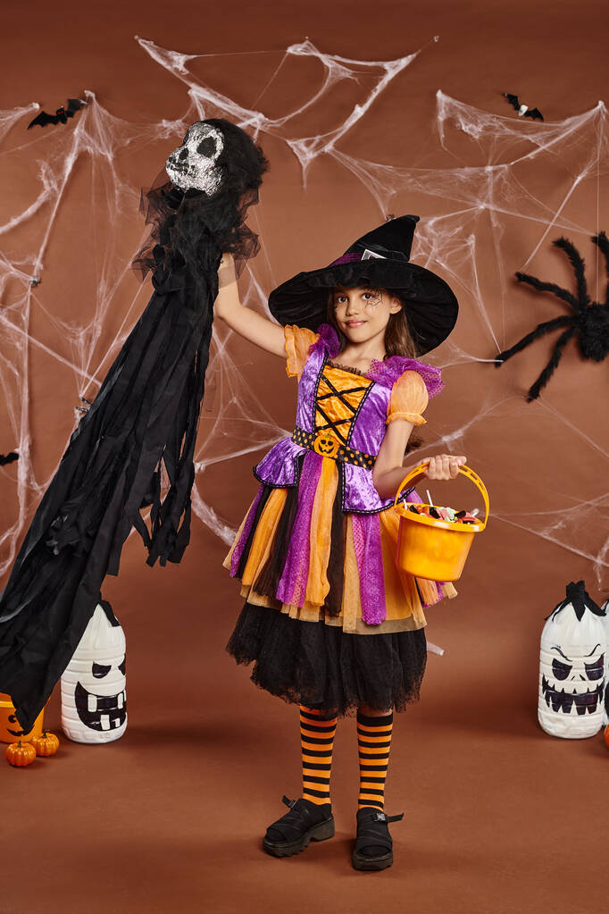 gelukkig kind in heks hoed met spinnenweb make-up houden emmer van snoep en spookachtig speelgoed, Halloween - Foto, afbeelding