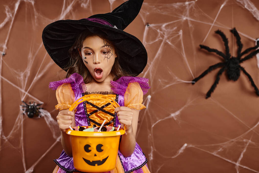 geschokt meisje in heks hoed met spinnenweb make-up op zoek naar snoep in Halloween emmer op bruin - Foto, afbeelding