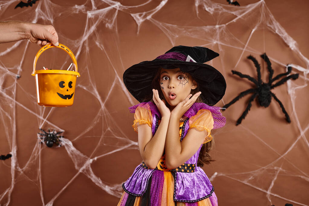 zszokowany dzieciak w kapeluszu czarownicy i kostium Halloween patrząc na kamerę w pobliżu strony trzymając słodycze w wiadrze - Zdjęcie, obraz
