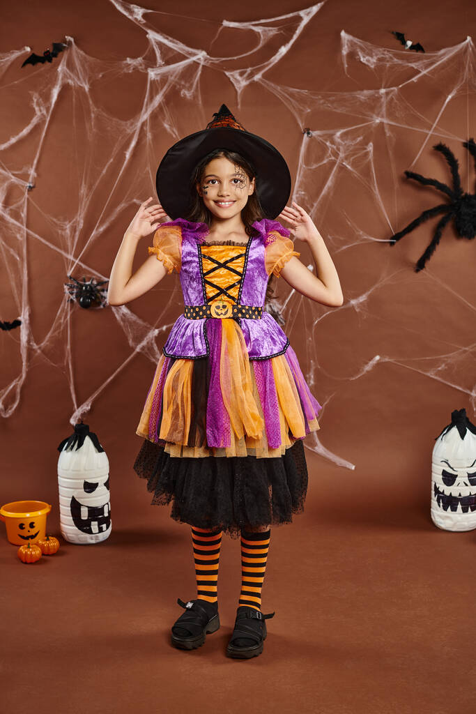 menina feliz no traje de Halloween ajustando chapéu de bruxa e de pé perto de teias de aranha no fundo marrom - Foto, Imagem