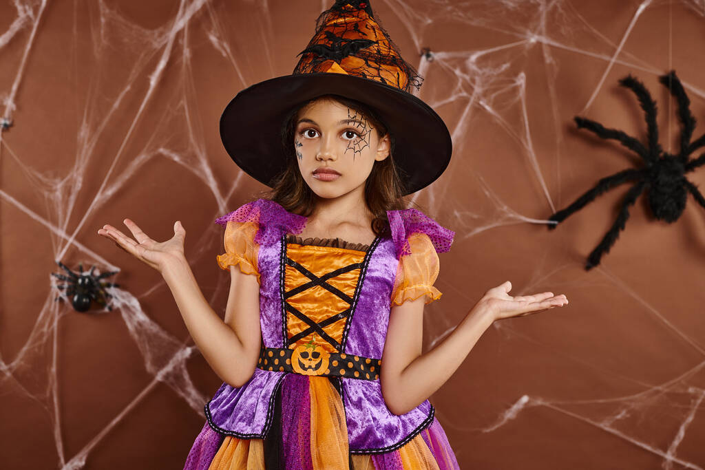 confused girl in witch hat and Halloween costume near cobwebs on brown backdrop, spooky season - Photo, Image