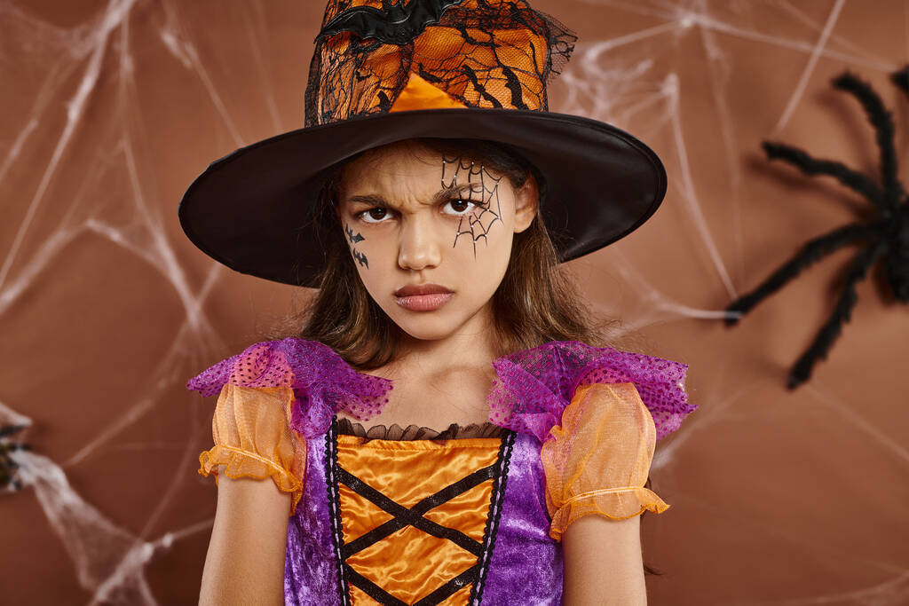 displeased girl in witch hat and Halloween costume frowning on brown background, spooky season - Photo, Image