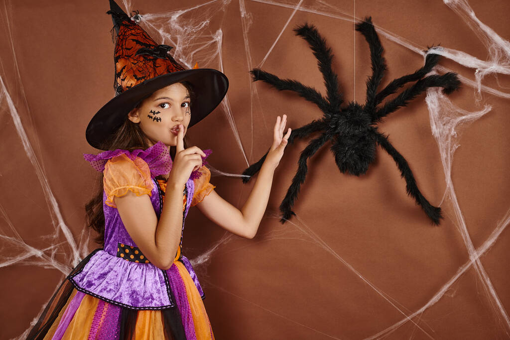 chica espeluznante en sombrero de bruja y disfraz de Halloween mostrando silencio cerca de araña falsa sobre fondo marrón - Foto, Imagen