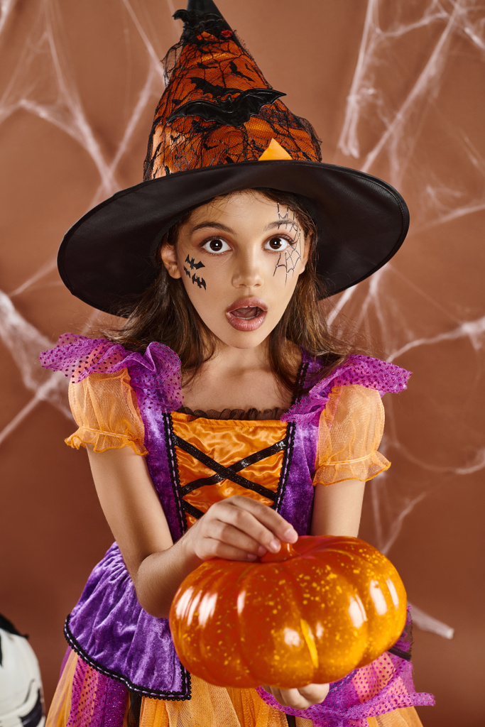 ragazza stupita in cappello da strega e costume di Halloween in piedi con zucca su sfondo marrone, ragnatele - Foto, immagini