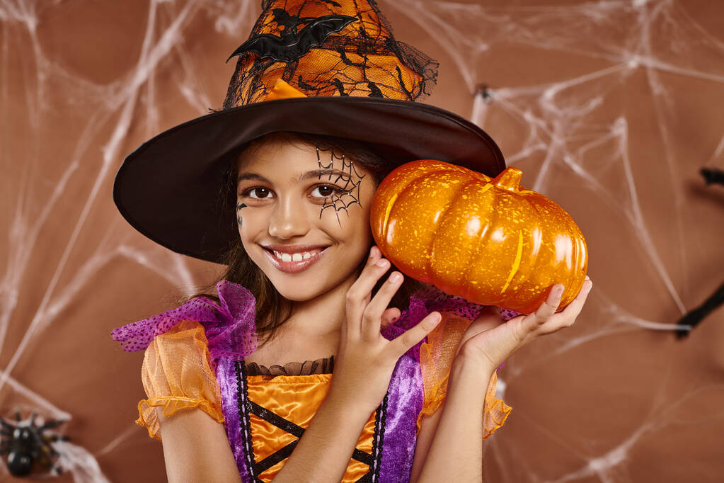 menina alegre em chapéu de bruxa e traje de Halloween em pé com abóbora no fundo marrom, teias de aranha - Foto, Imagem