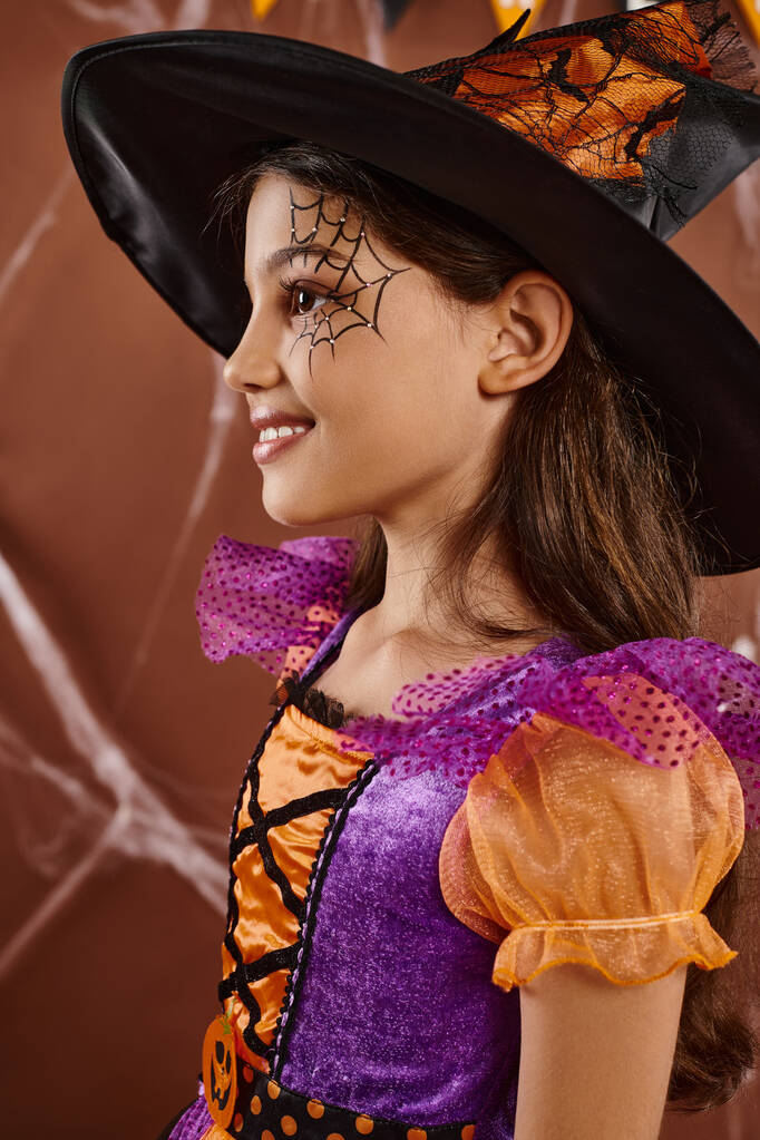 schattig klein meisje in Halloween heks kostuum en puntige hoed glimlachen op bruine achtergrond, zijaanzicht - Foto, afbeelding
