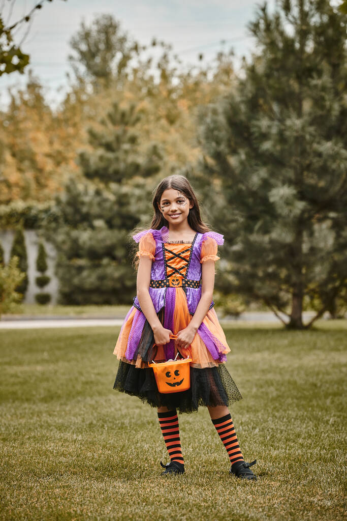 cute child in Halloween costume standing with bucket of sweet candies on green grass, girl in dress - Photo, Image