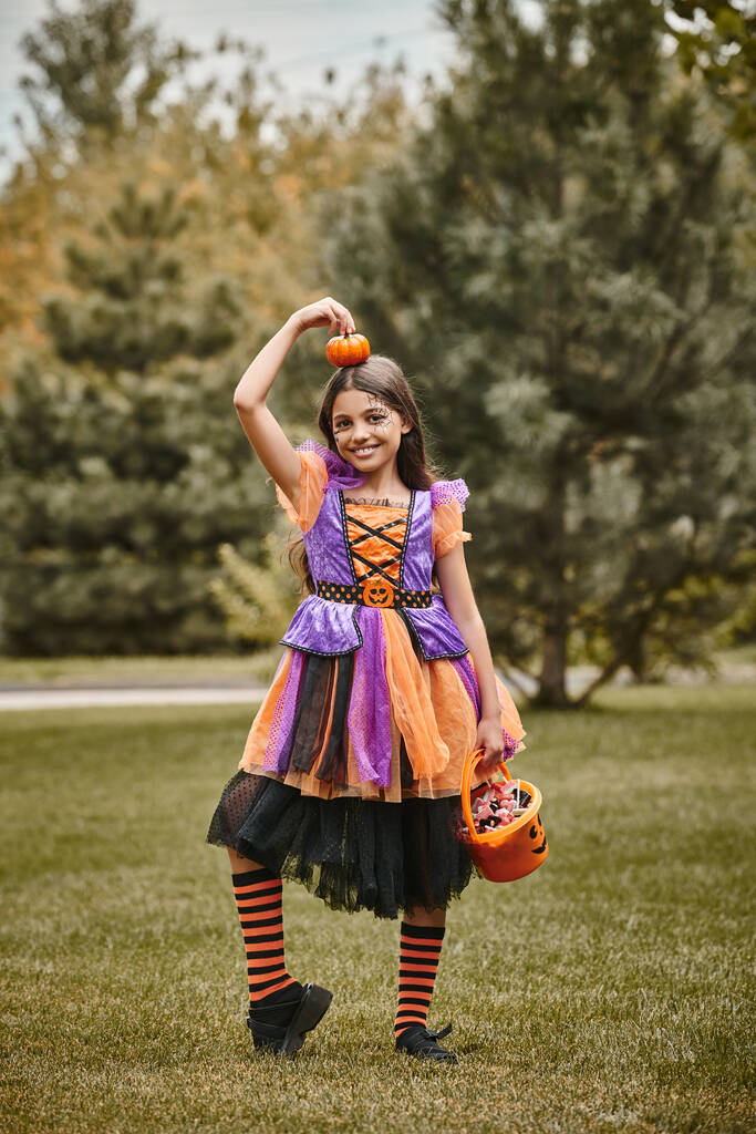 happy girl in Halloween costume holding pumpkin and bucket of candies on green grass, kid in dress - Photo, Image