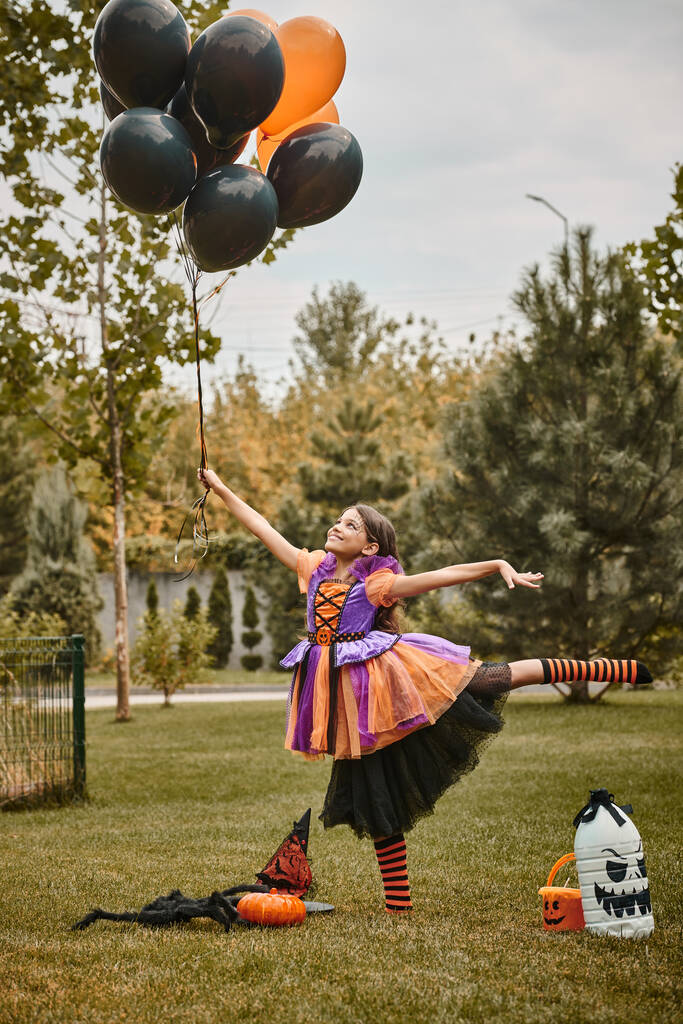 cheerful girl in Halloween attire holding balloons near pumpkin, witch hat and candy bucket on grass - Photo, Image