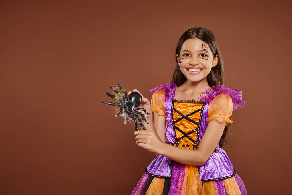 joyous girl in Halloween costume with spiderweb makeup holding fake spider on brown backdrop, spooky - Photo, Image