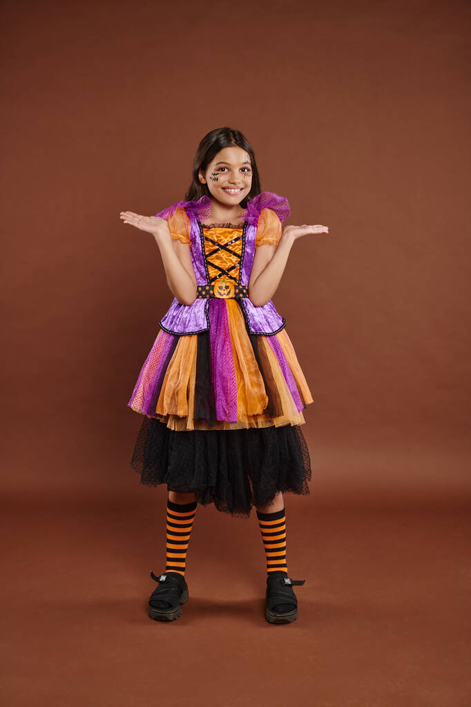 chica divertida en disfraz de Halloween con maquillaje spiderweb sonriendo y haciendo gestos en el fondo marrón - Foto, imagen