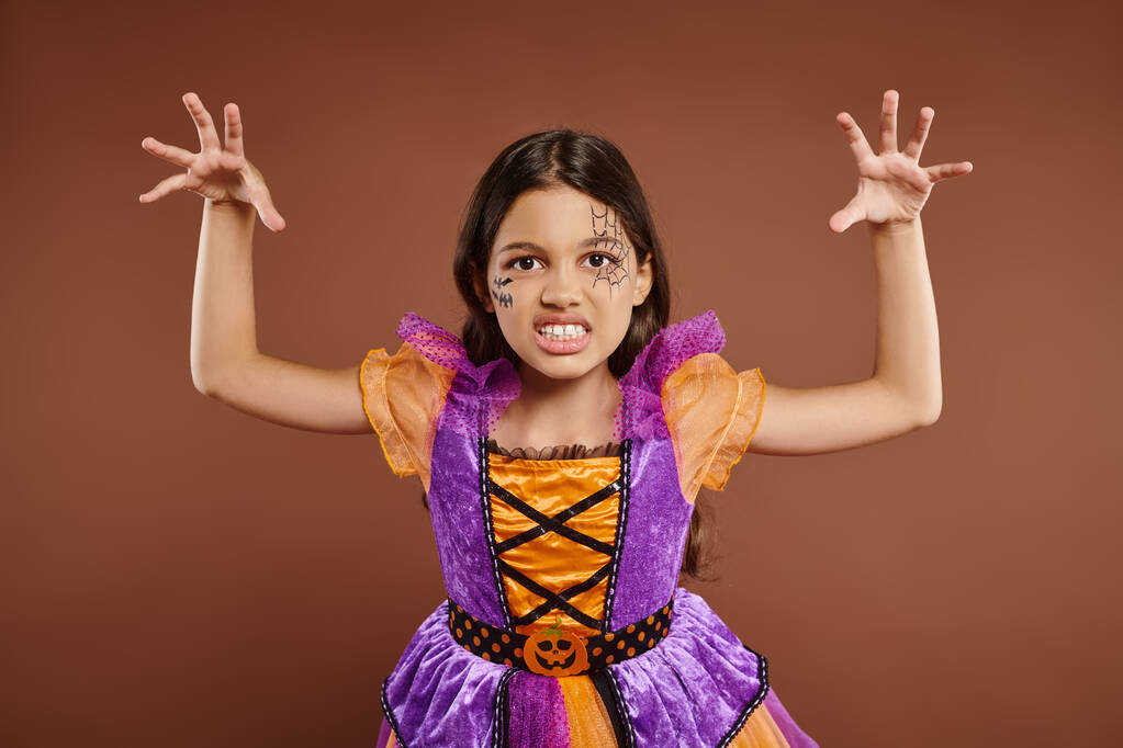 жуткий ребенок в костюме Хэллоуина с паутиной-макияжем рычит и жестикулирует на коричневом фоне - Фото, изображение